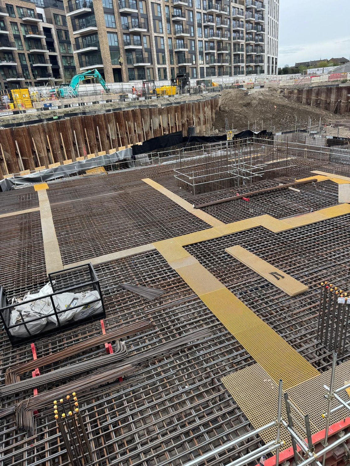 Deep basement excavation with sheet-piled retaining wall, excavator and reinforcement to the base, with tower blocks under construction behind